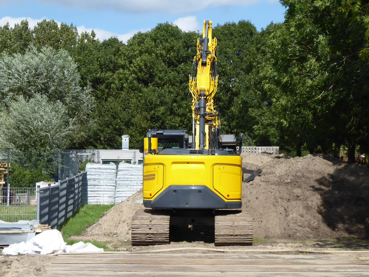 Yellow tracked excavator parked beside soil piles on a cleared lot at the edge of the tree line