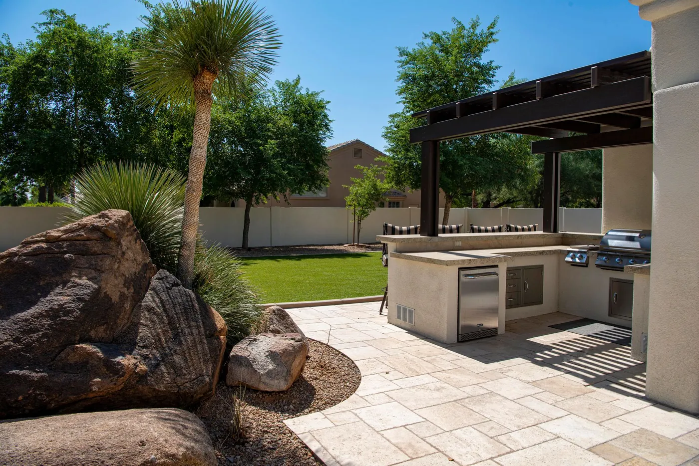 Paver patio with a built-in outdoor kitchen under a pergola, edged by boulders and drought-tolerant plantings