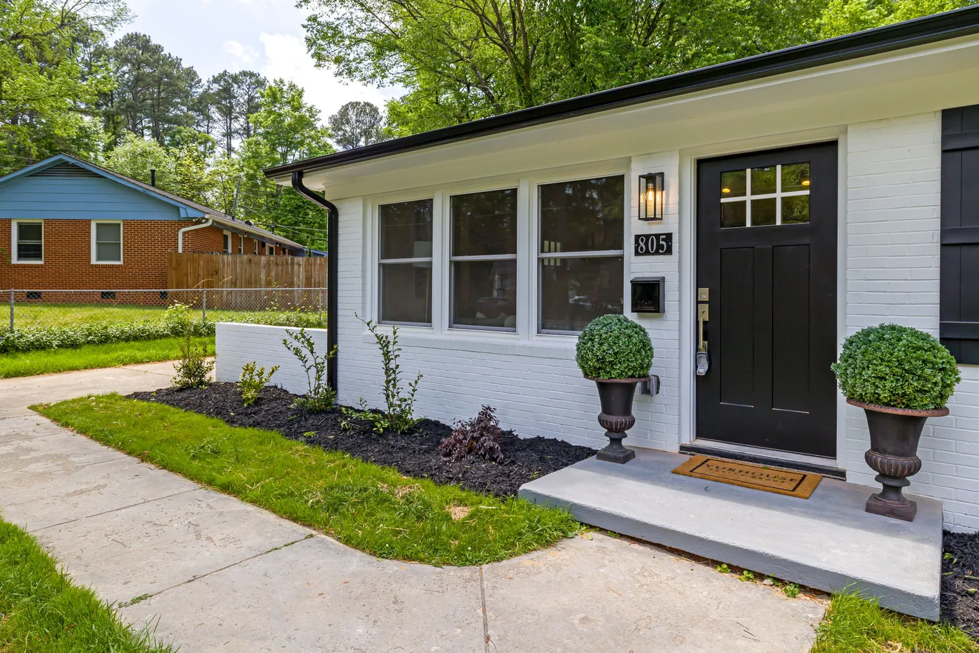 Newly planted front entry beds with low boxwood shrubs, dark mulch and matching urn planters beside a white house