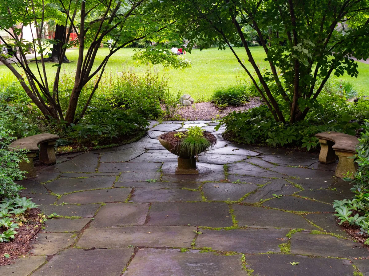 Flagstone patio with stone benches and a planted urn, framed by shade trees and garden beds